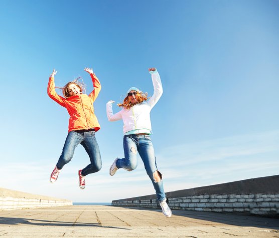 Two smilng women jumping in the air on a stone sea wall with the sea in the background.